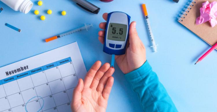 There is a person with a glucose monitor in their right hand, their left hand is out to prick their finger. Their hands are resting on a blue background. Around their hands are pens, pencils, an apple, journal and calendar on the November page.