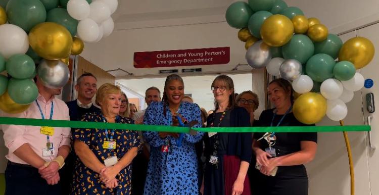 North West London employees are standing in a group behind a green ribbon. There is a lady in a blue address who has scissors to cut the ribbon.