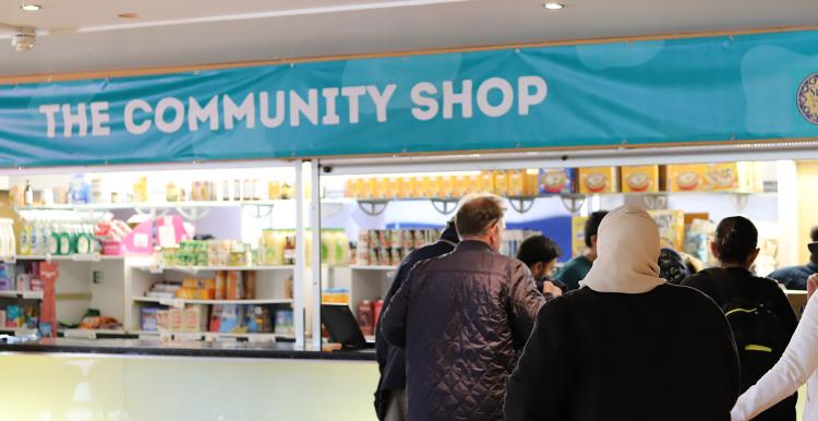 There is a large blue banner with the words 'The community shop' written in white capital letters. There is a staff member on the left, and service users are queuing up on the right hand side behind the counter. The middle has multiple shelves of food and other products.