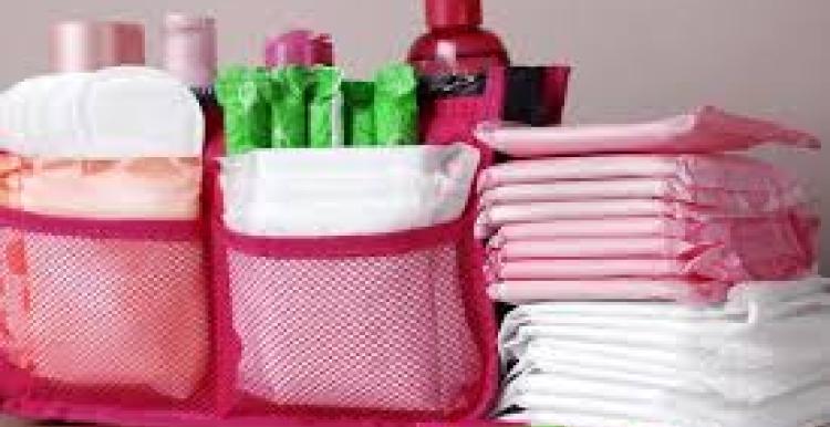 There are three piles of period products. Two piles have period pads in a pink mesh pouch. The third pile is pads stacked on top of each other, the pads are pink and white. There is a purple, green and blue tampon infront of the stack.