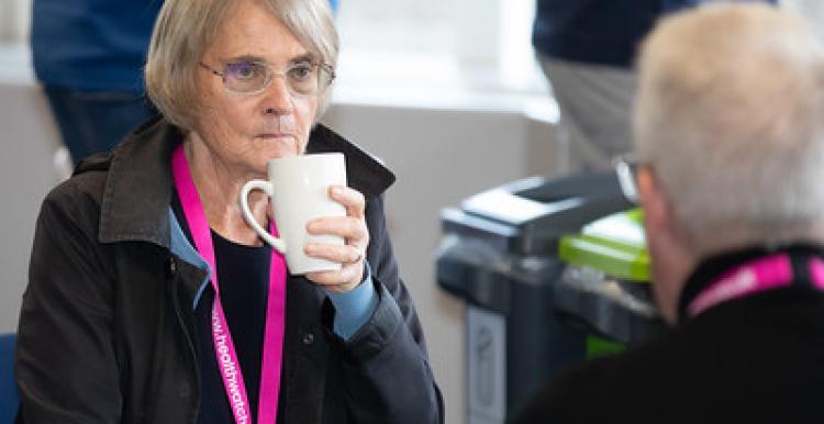 On the left is an elderly lady who has white hair, glasses and she is wearing a black top and black jacket. She is looking at a man infront of her while holding a coffee cup. The elderly man infront of her is also wearing a black top and Healthwatch lanyard like her.