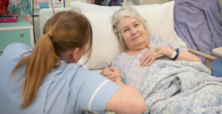 There is a white-haired elderly woman lying down on a hospital bed. Her left hand is upon her chest and her right hand is holding the hand of a female healthcare professional. The professional is a brown haired lady who is facing the elderly lady, so the audience cannot see her face. The professional has her hair in a ponytail with a strand next to her temples and she is wearing light blue scrubs.