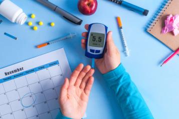 There is a person with a glucose monitor in their right hand, their left hand is out to prick their finger. Their hands are resting on a blue background. Around their hands are pens, pencils, an apple, journal and calendar on the November page.