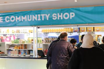 There is a large blue banner with the words 'The community shop' written in white capital letters. There is a staff member on the left, and service users are queuing up on the right hand side behind the counter. The middle has multiple shelves of food and other products.