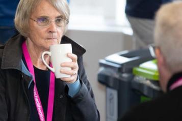 On the left is an elderly lady who has white hair, glasses and she is wearing a black top and black jacket. She is looking at a man infront of her while holding a coffee cup. The elderly man infront of her is also wearing a black top and Healthwatch lanyard like her.