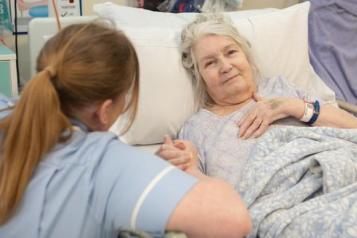 There is a white-haired elderly woman lying down on a hospital bed. Her left hand is upon her chest and her right hand is holding the hand of a female healthcare professional. The professional is a brown haired lady who is facing the elderly lady, so the audience cannot see her face. The professional has her hair in a ponytail with a strand next to her temples and she is wearing light blue scrubs.