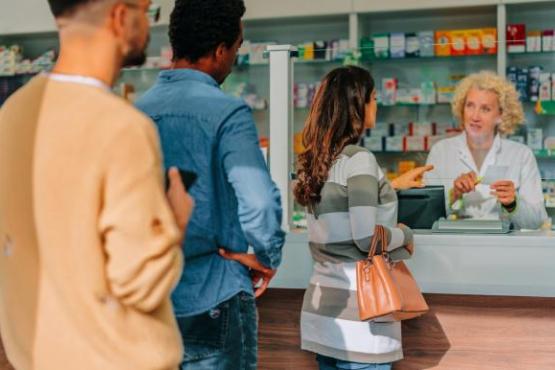 This is a scene of a pharmacy. The pharmacist has white skin, short curly blonde hair and is wearing a white lab coat. She is behind a white counter. There are three patients in the queue in front of the counter. The first patient is a woman with brown curly hair, checkered coat and is holding a handbag on her arm. The man behind her has black hair and is wearing a denim outfit. The final patient is a bearded man with brown hair and wearing a yellow jumper.