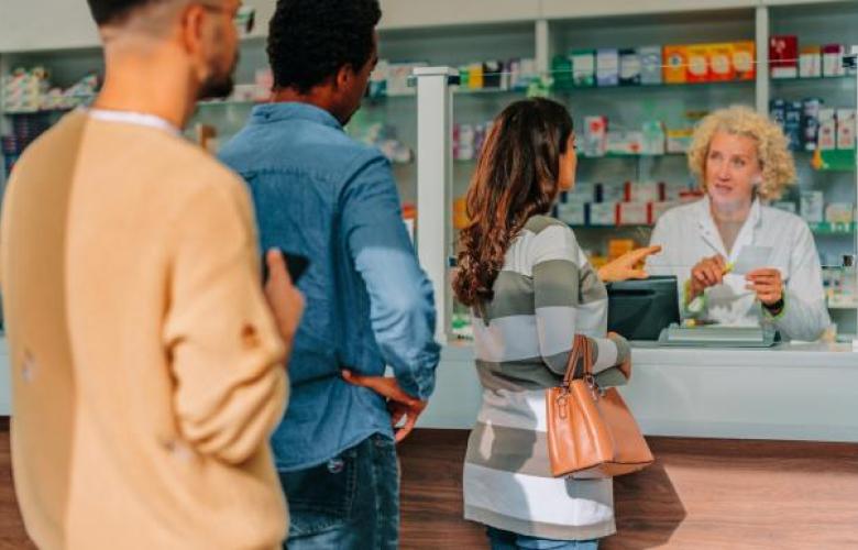 This is a scene of a pharmacy. The pharmacist has white skin, short curly blonde hair and is wearing a white lab coat. She is behind a white counter. There are three patients in the queue in front of the counter. The first patient is a woman with brown curly hair, checkered coat and is holding a handbag on her arm. The man behind her has black hair and is wearing a denim outfit. The final patient is a bearded man with brown hair and wearing a yellow jumper.