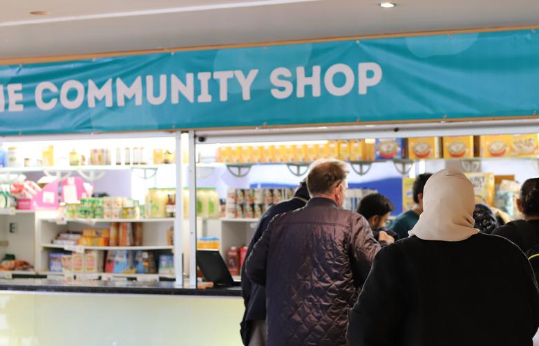 There is a large blue banner with the words 'The community shop' written in white capital letters. There is a staff member on the left, and service users are queuing up on the right hand side behind the counter. The middle has multiple shelves of food and other products.