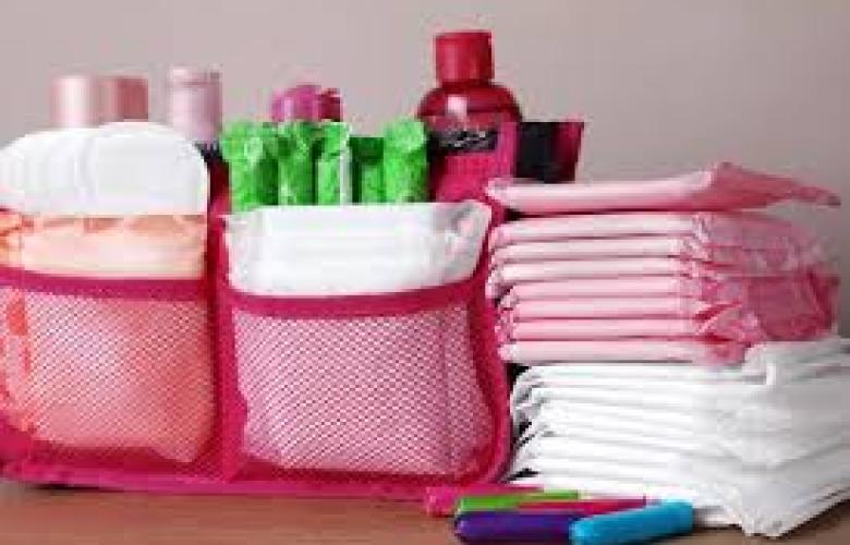 There are three piles of period products. Two piles have period pads in a pink mesh pouch. The third pile is pads stacked on top of each other, the pads are pink and white. There is a purple, green and blue tampon infront of the stack.