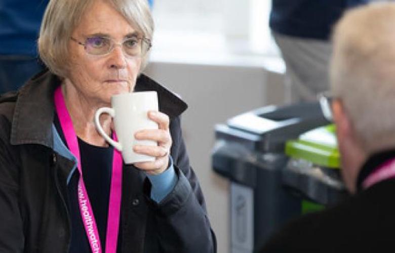 On the left is an elderly lady who has white hair, glasses and she is wearing a black top and black jacket. She is looking at a man infront of her while holding a coffee cup. The elderly man infront of her is also wearing a black top and Healthwatch lanyard like her.