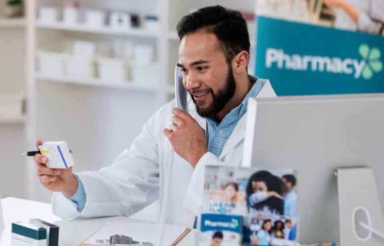 There is a male pharmacist who has dark brown hair and a beard. He is wearing a blue button up shirt with a lab coat on top of that. He is holding a white packet of tablets. On the right hand side there is a Macbook.