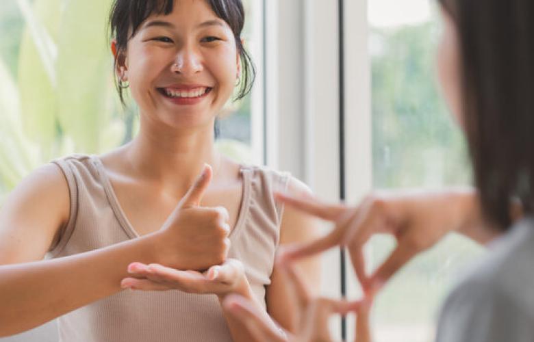 There is one lady in this picture. On the right hand side corner, we can see her shoulder and left side of her hair as she is facing a mirror. In the middle of the photo, there is a woman who has black hair with a fringe and she is smiling while practicing British Sign Language