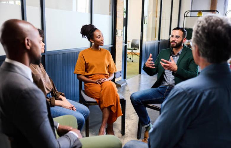 This photo is showing a focus group and there are four people. There is one man on the left hand side and one man on the right hand side, both of them are facing two other people in front. So the faces of these men cannot be seen. In the middle of the photo is a lady wearing an orange dress and a man sitting beside her, talking to the group. The men are wearing smart suits.