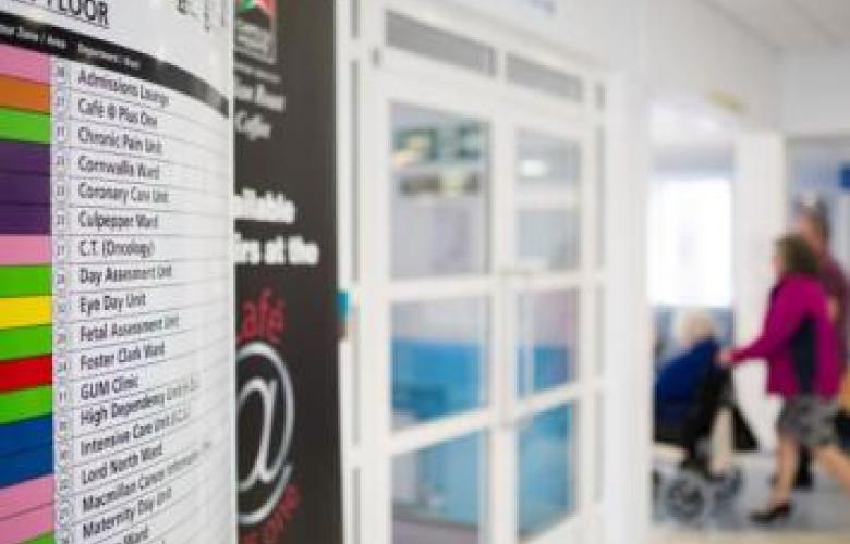 This is a picture of a hospital floor. On the left hand side there is a list of different rooms within the floor. The middle has white square windows and the right hand side has a lady pushing an individual in a wheelchair into the room.